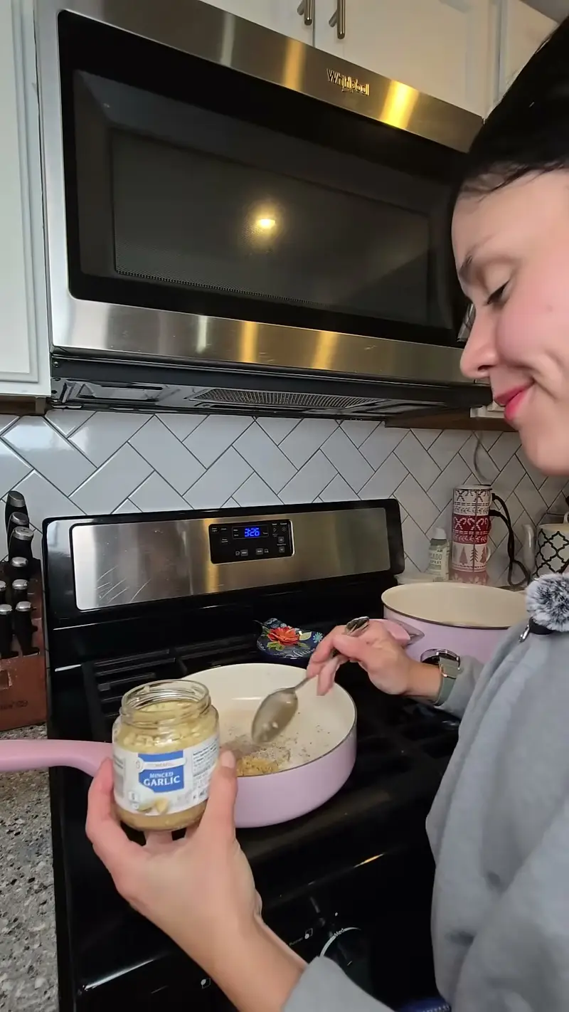 Crystal spooning minced garlic from a jar into a pink skillet on the stove