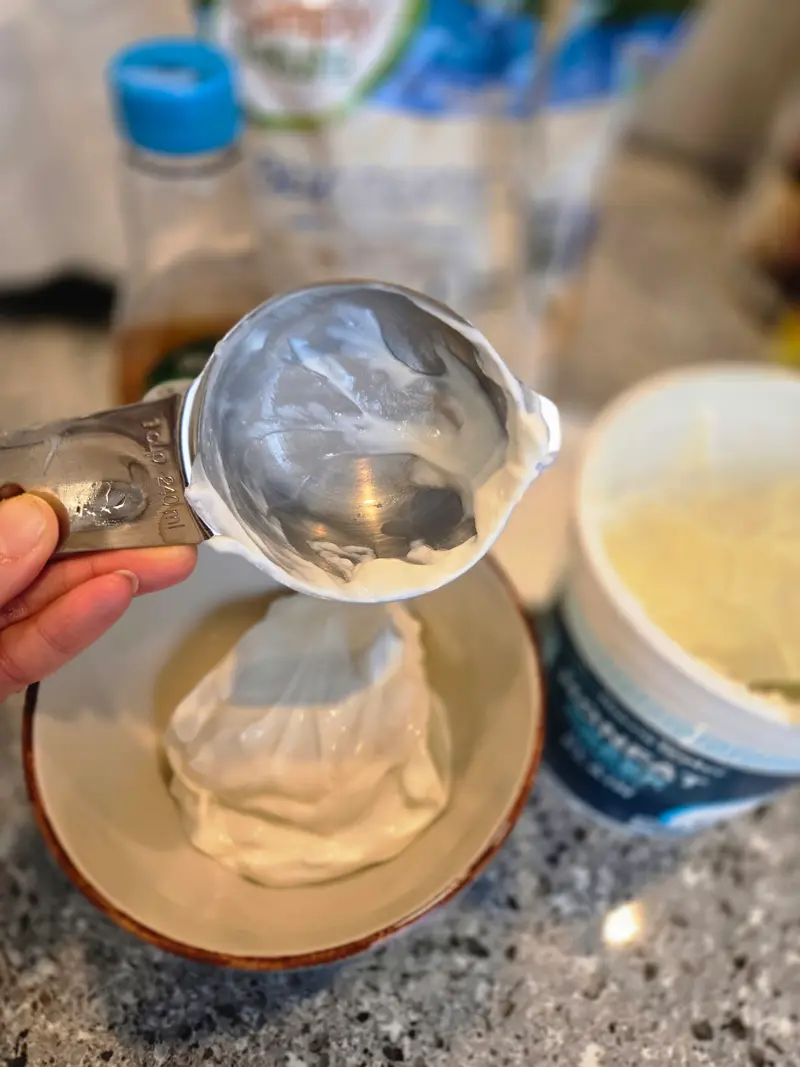 Scooping Greek yogurt into a small bowl with a measuring cup