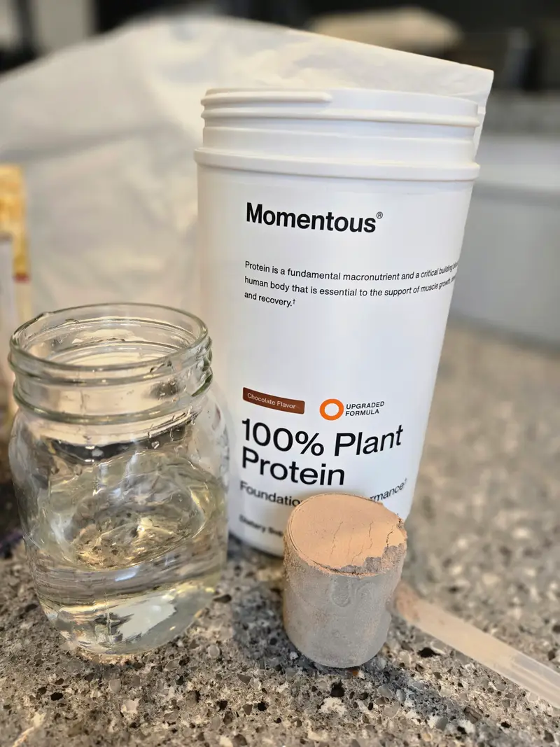 An empty glass jar beside a tub of Momentous plant protein and a full scoop of powder