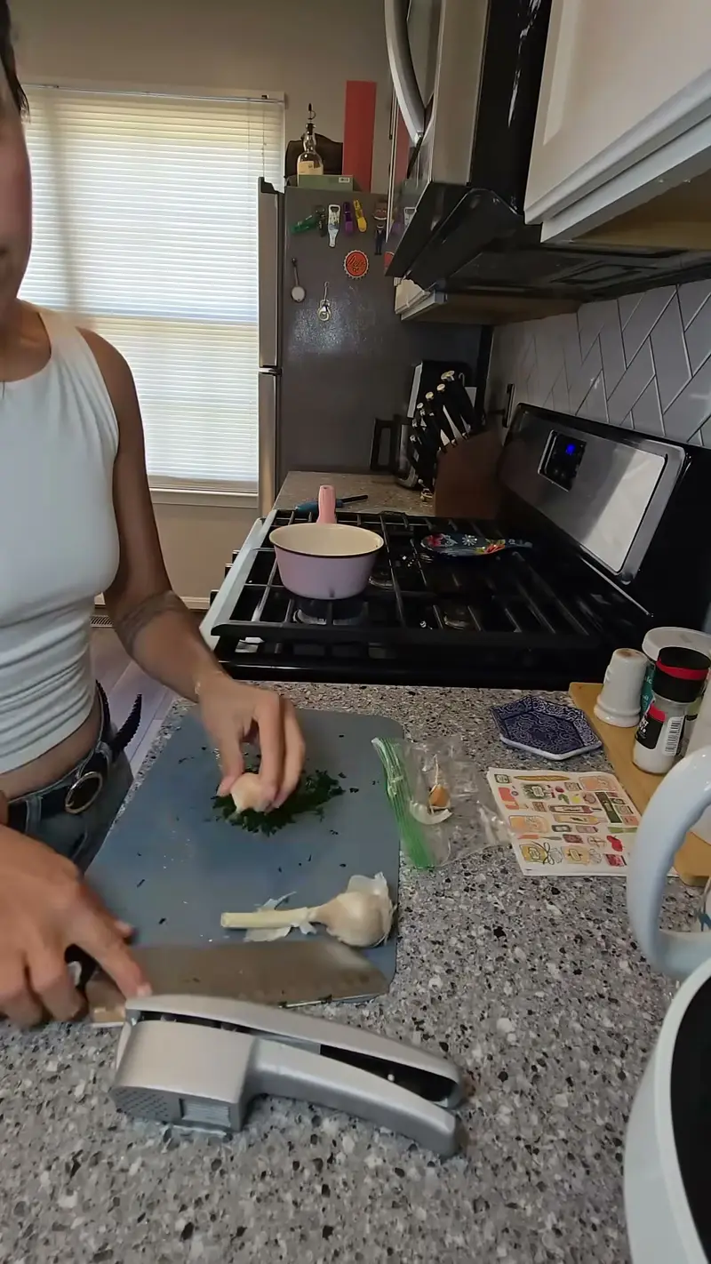 Crystal crushing garlic with a press next to chopped herbs on a board